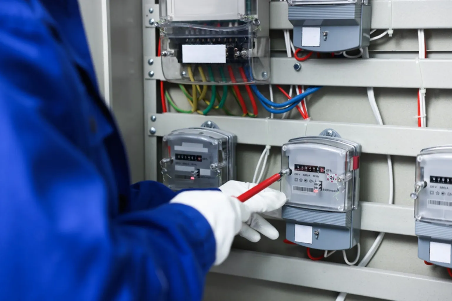 Elektriker i blå arbejdsjakke og hvide handsker kontrollerer elmålere i et måleskab