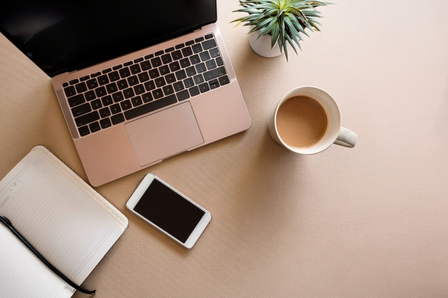 En bærbar computer, en mobiltelefon, en notesbog og en kop kaffe på et lyst skrivebord set ovenfra