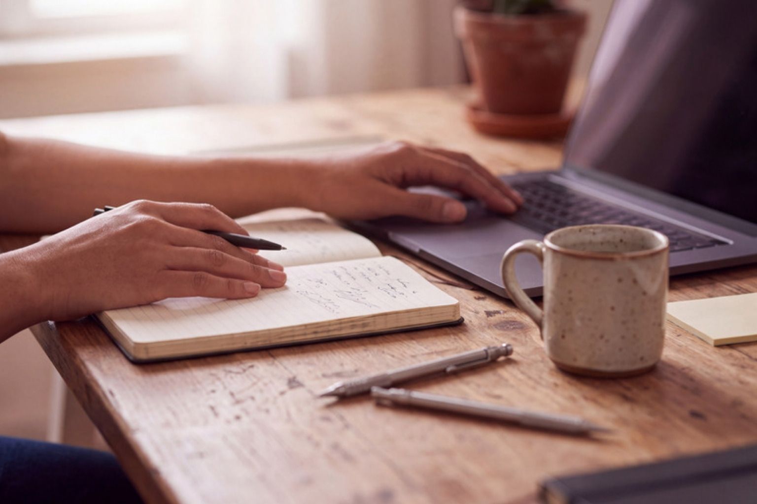 En person skriver noter i en notesbog ved et træbord med bærbar computer og kaffekrus